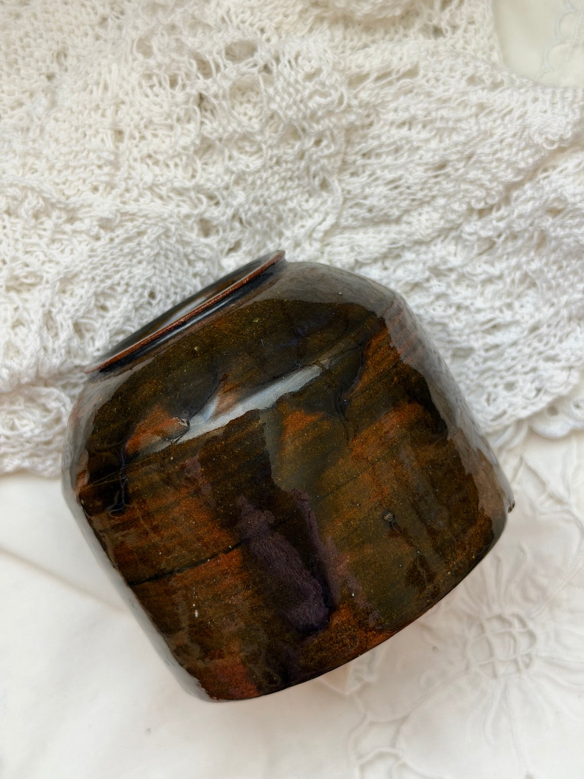 A small handmade ceramic pot with a glossy, marbled finish in dark brown, black, and amber tones, resting on a white lace textile background.